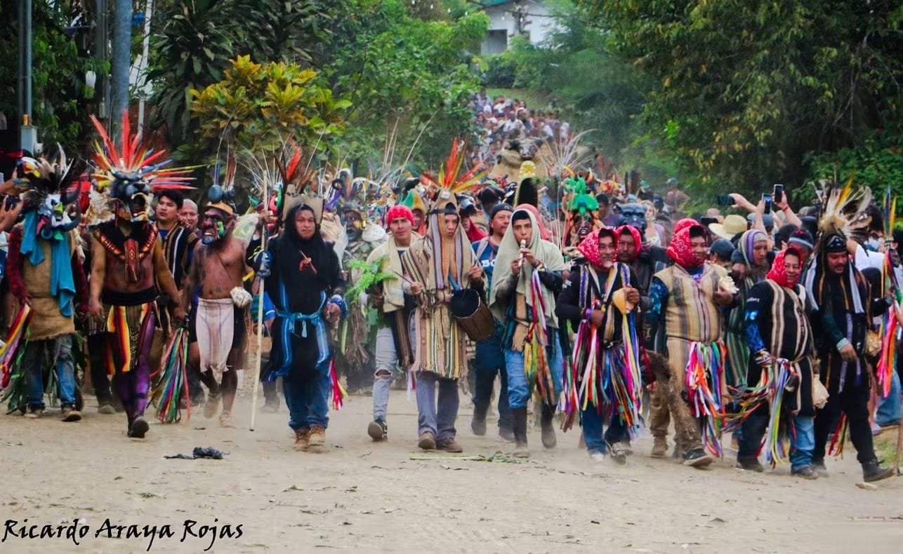Procesión de guerreros, músicos y comunidad avanzando por un camino de tierra rodeado de árboles